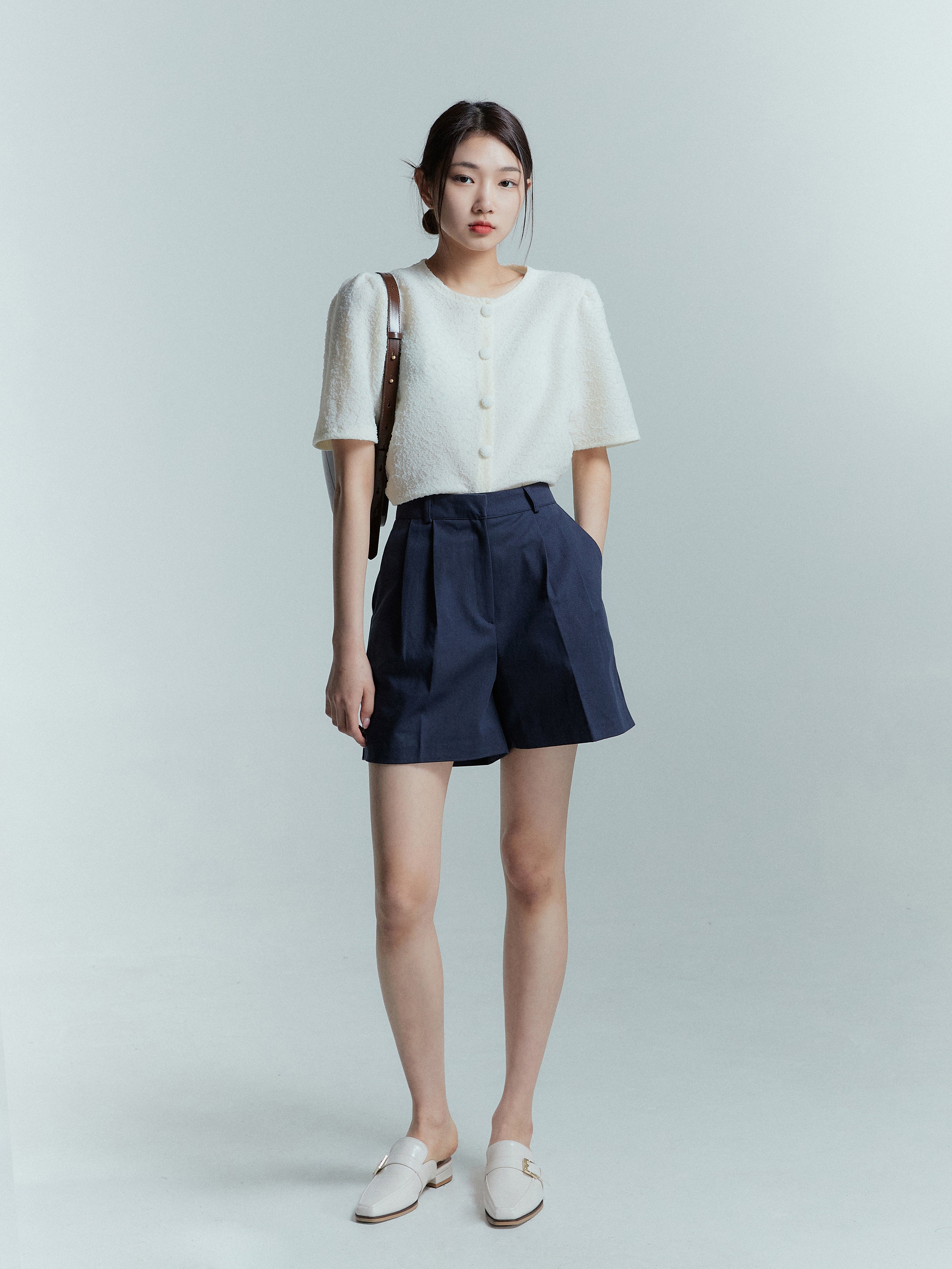 A full-length shot of a model wearing the navy pleated trouser shorts matched with ivory knit cardigan.