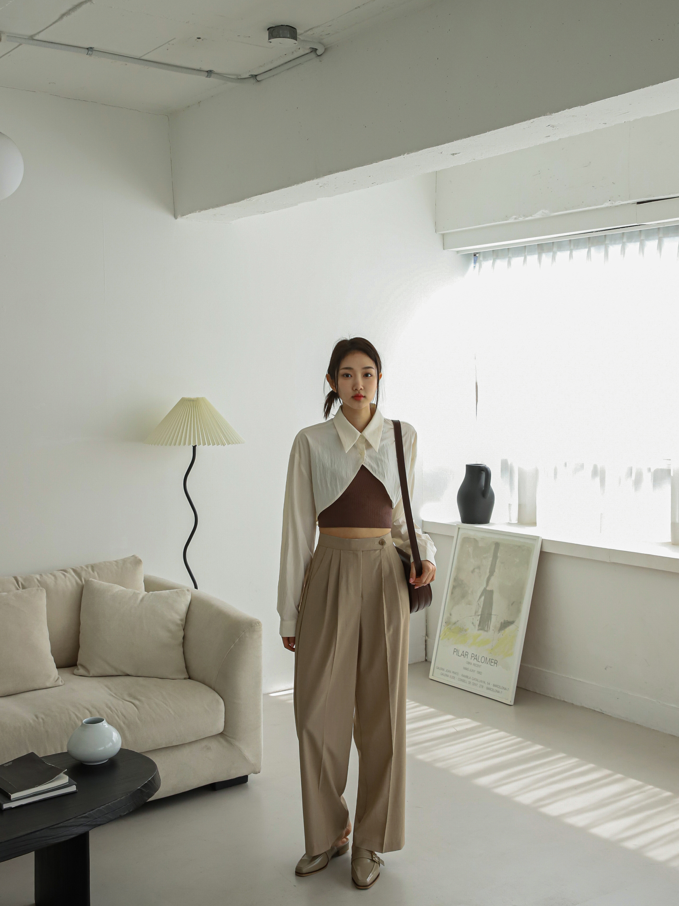 A long shot of a model standing straight wearing the beige button pleated trouser matching with the ivory cropped shirt. 