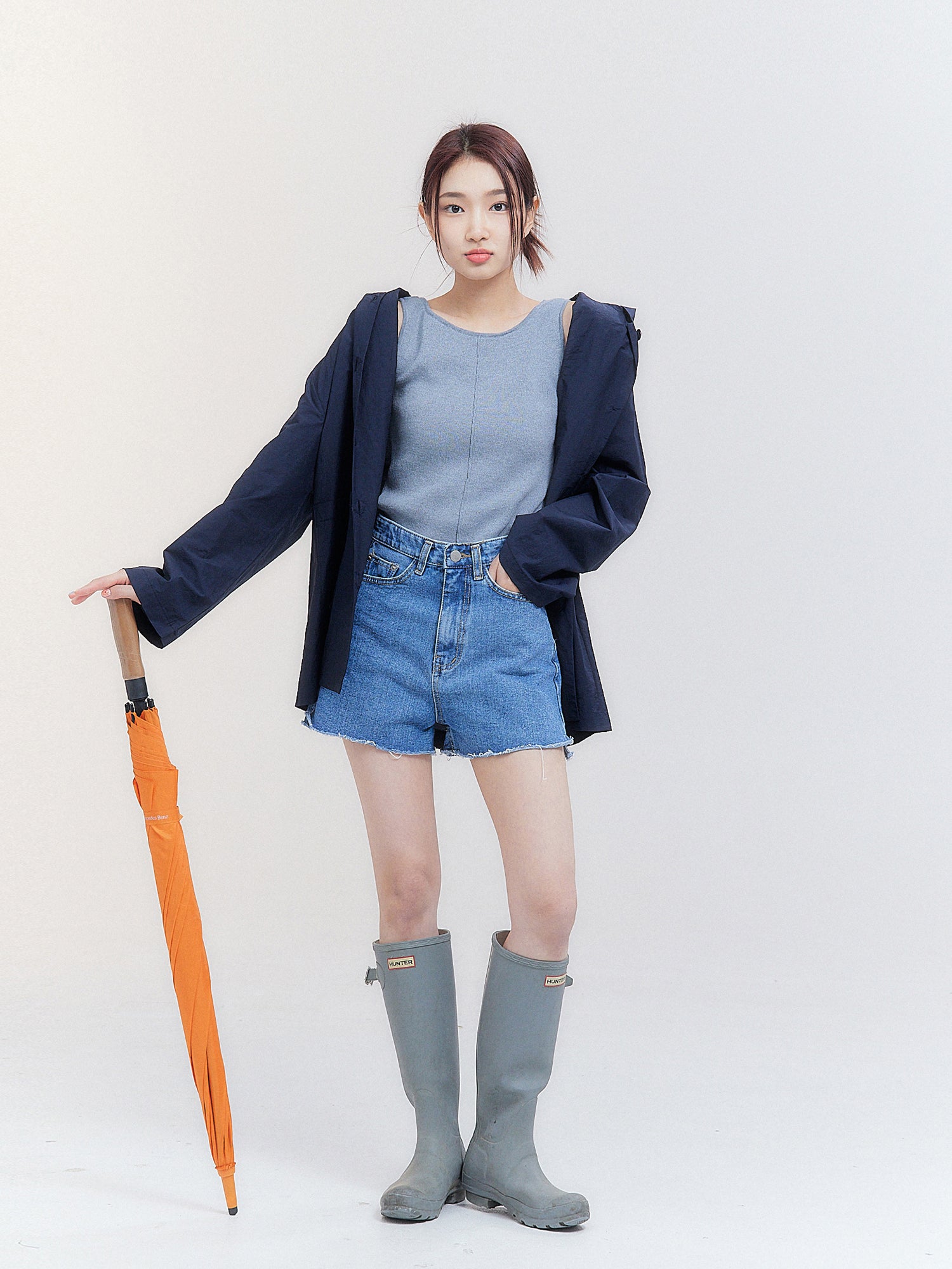 A full-length shot of the model wearing the hooded raincoat, skillfully styled with an orange umbrella, embodying the essence of Korean street fashion.