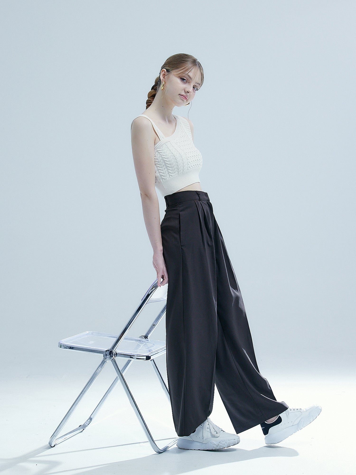 The model strikes a pose, leaning effortlessly against the chair, while sporting a fashionable ensemble featuring pleated pants.