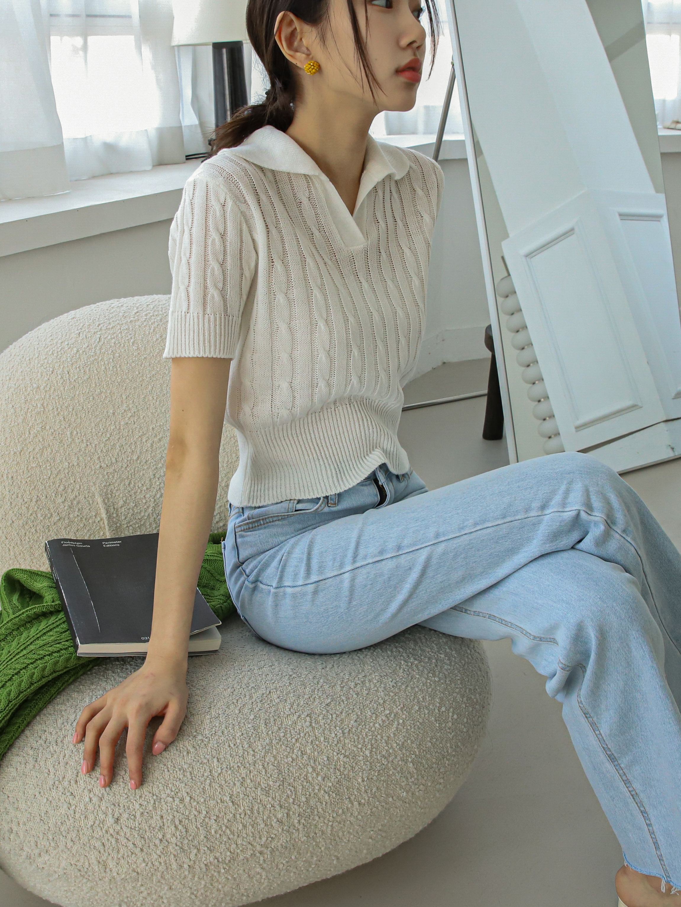 A picture of a model in a side angle seating on a sofa wearing the ivory knit shirt.