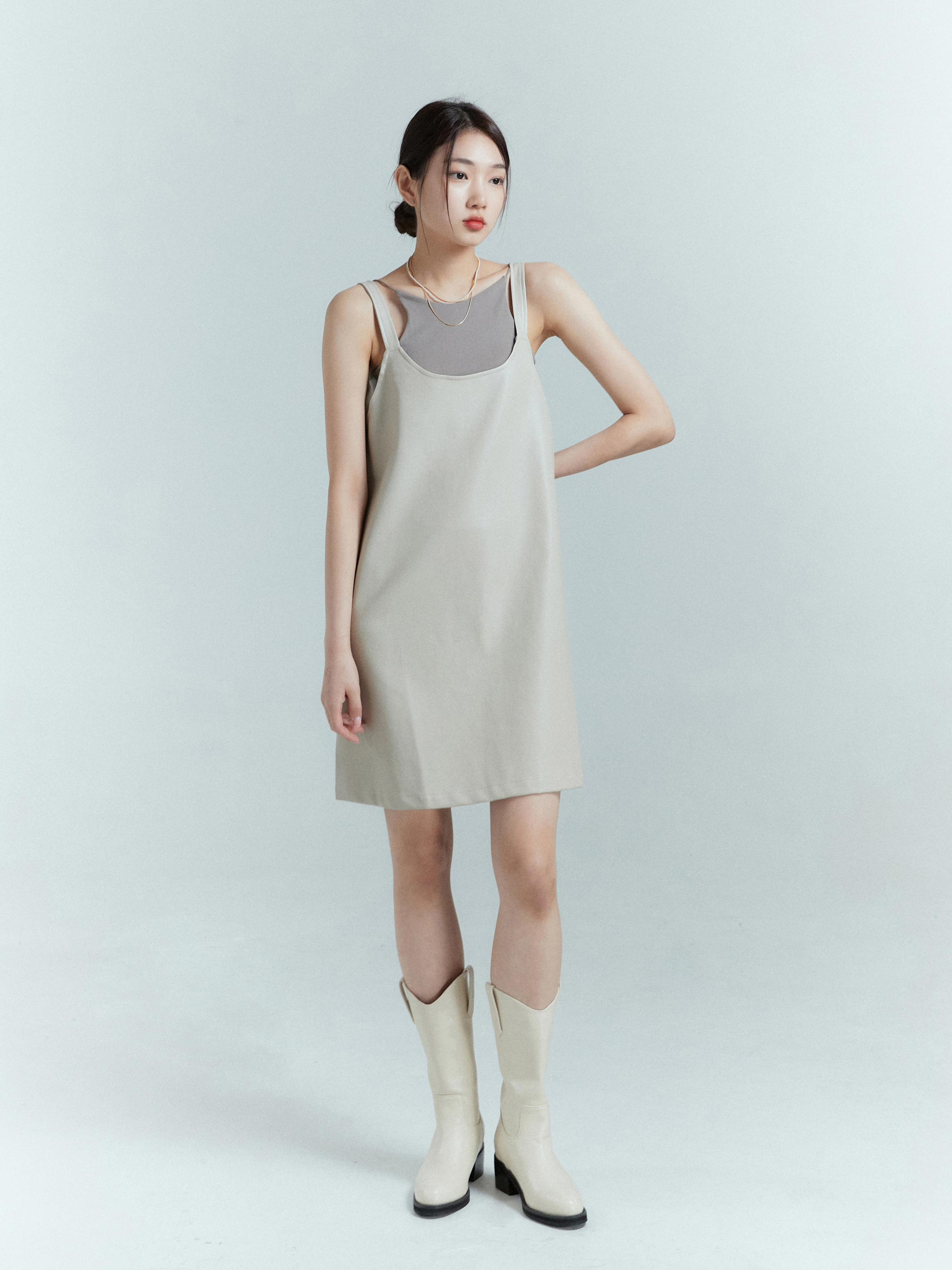  A stylish model gazes into the distance while dressed in a light beige tank dress and coordinating light beige boots, creating a fashionable look.