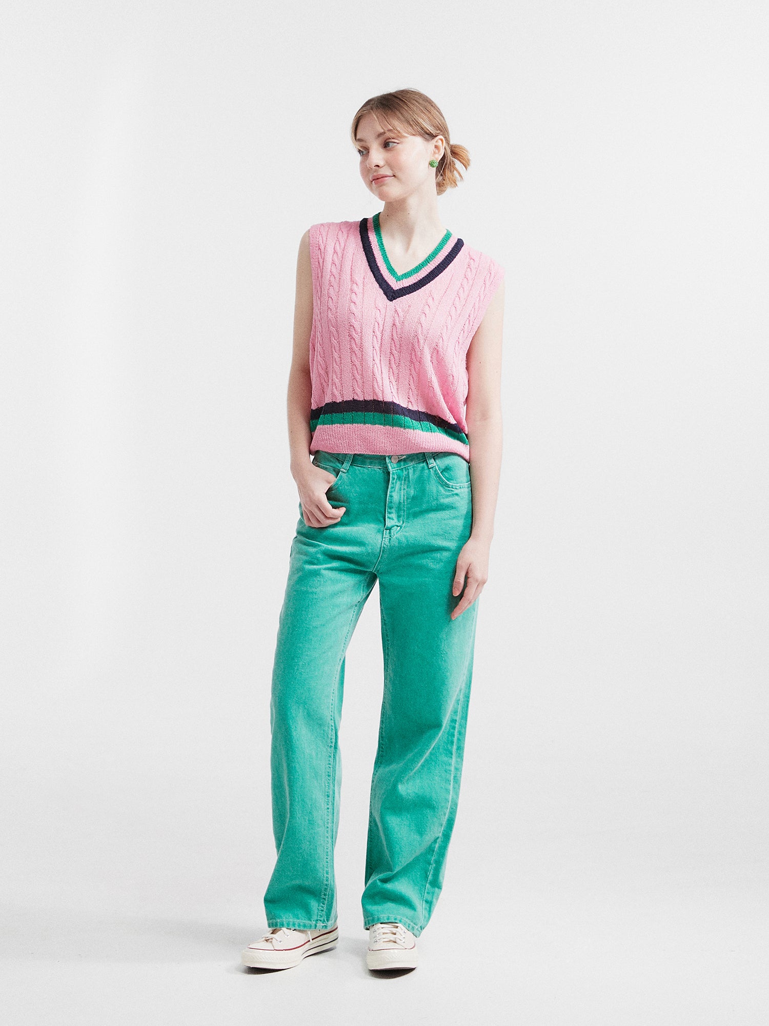 Model standing in contemporary fashion, wearing the pink V-neck knit vest, perfectly matching it with the ocean green carpenter jeans.