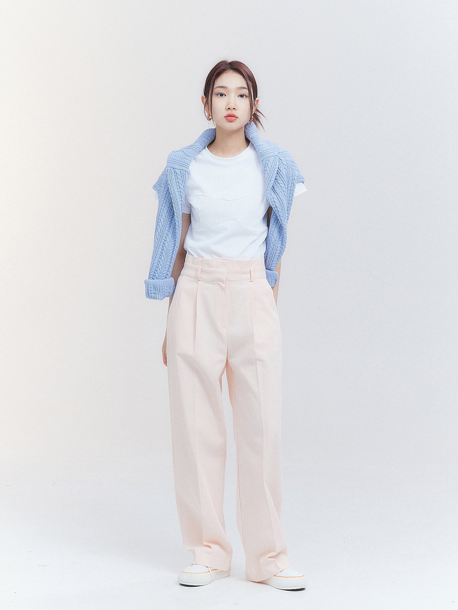 The image captures the model elegantly dressed in pink belted pleated trousers, complemented by a sweater draped over her shoulders.