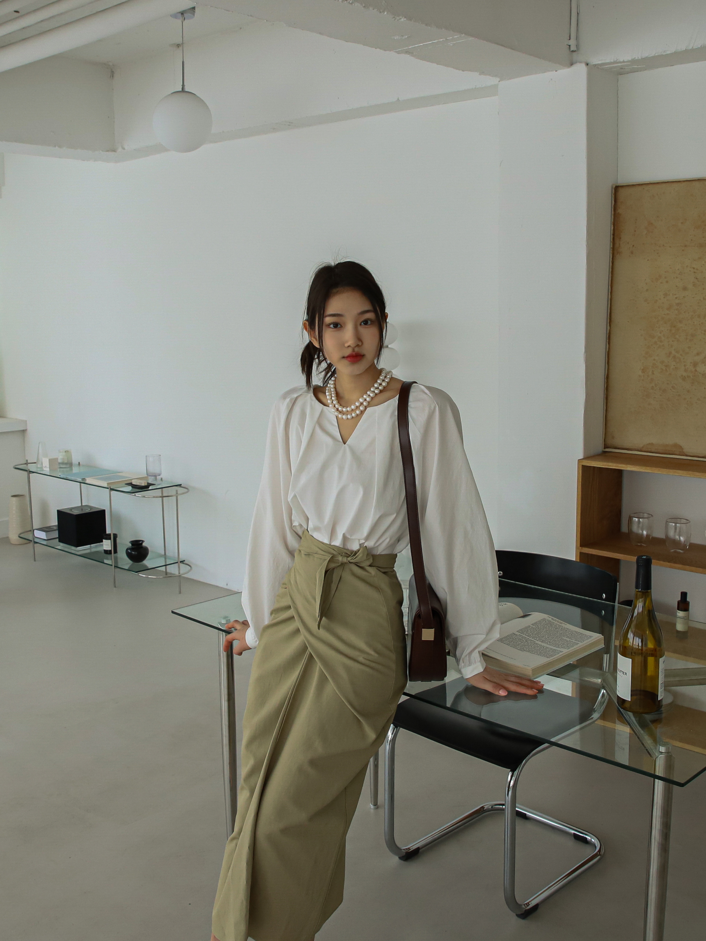 A tilted angle of a model standing against a glass table, showcasing the alluring beige wrap midi skirt.