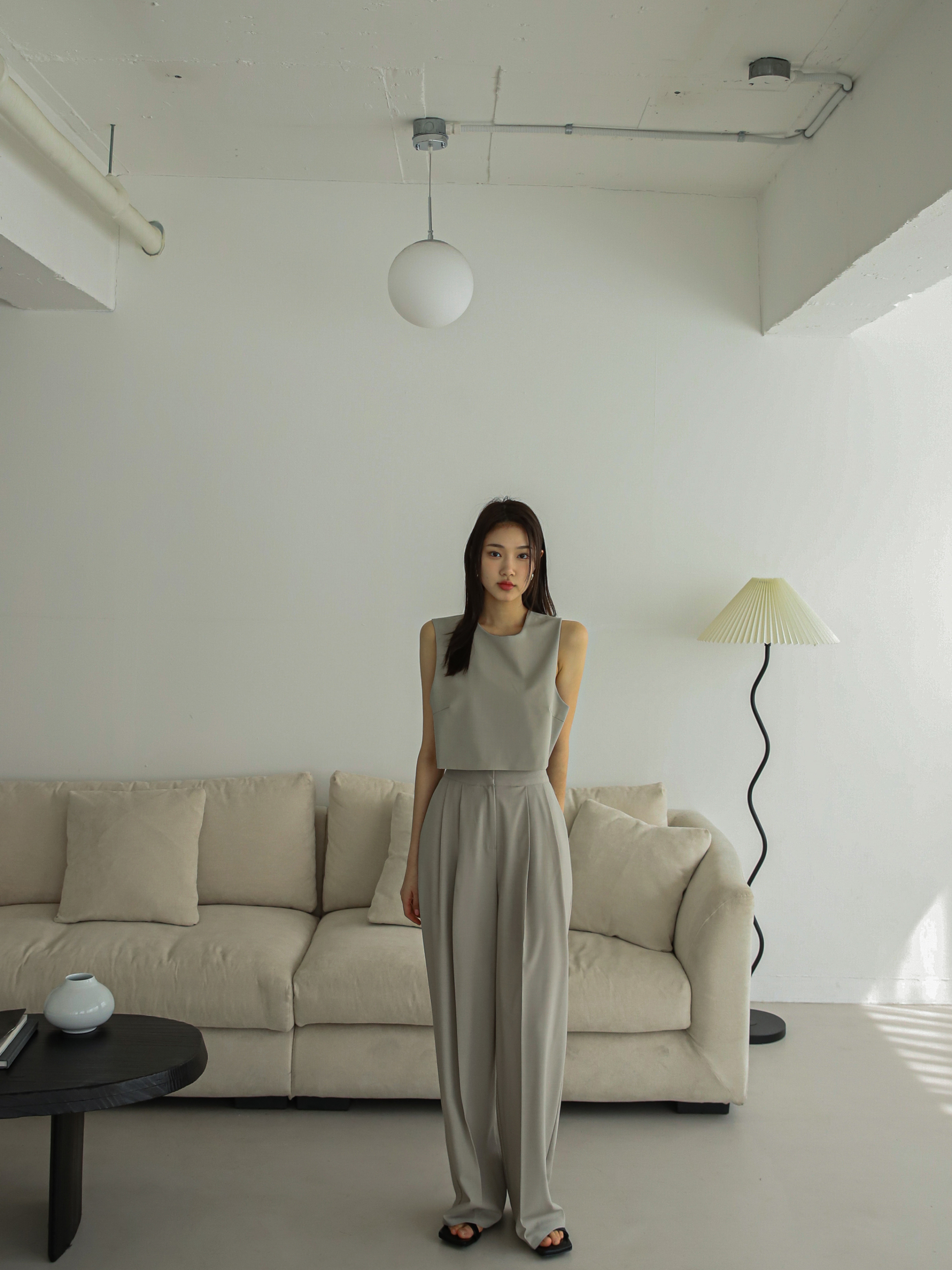 A confident model stands tall, gazing straight ahead, showcasing the beige sleeveless blazer top paired with pleated trousers in a captivating long shot.